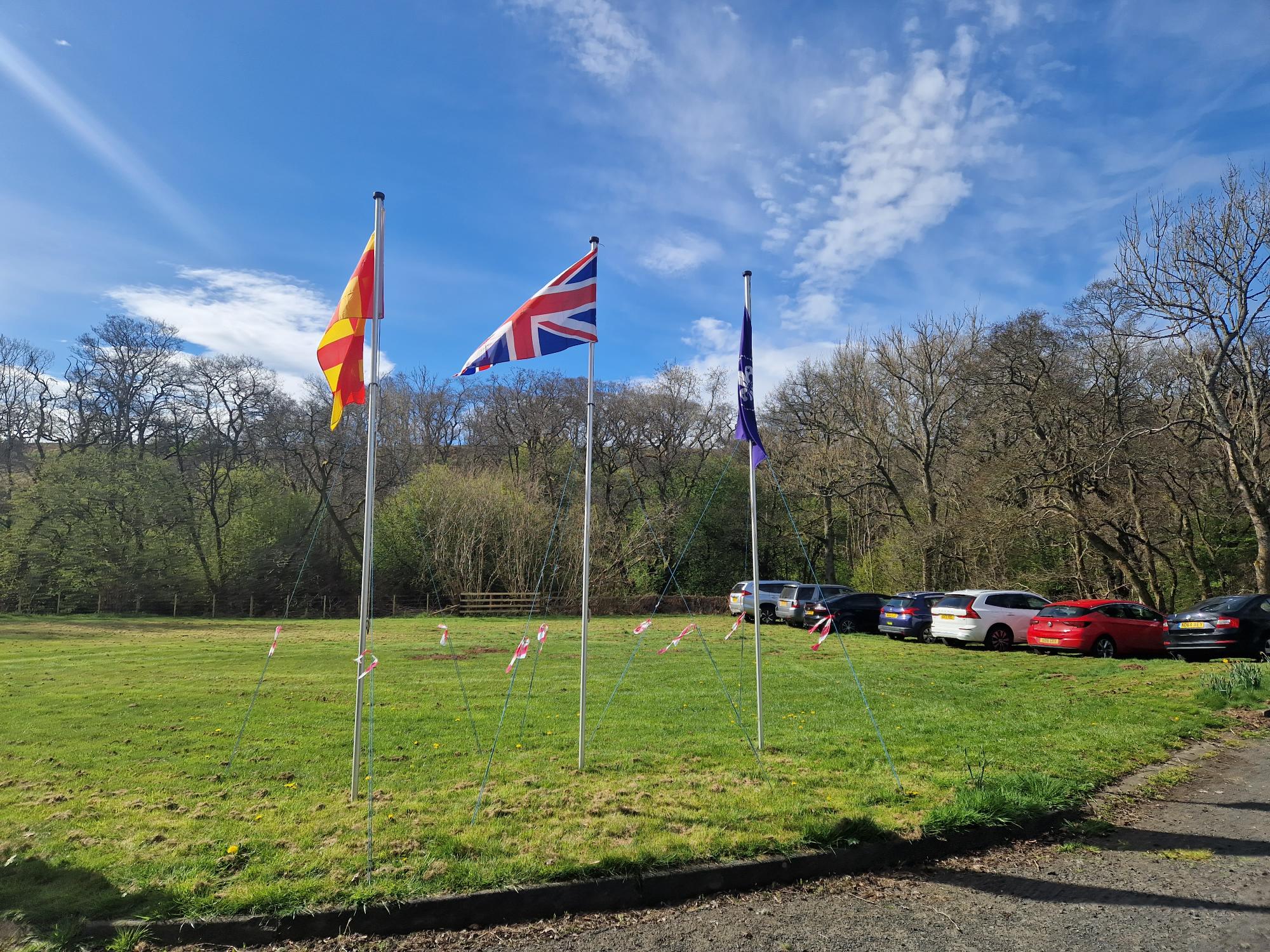 Photo of Nunnykirk site with three flag poles standing on a grassed area with woodland in the background