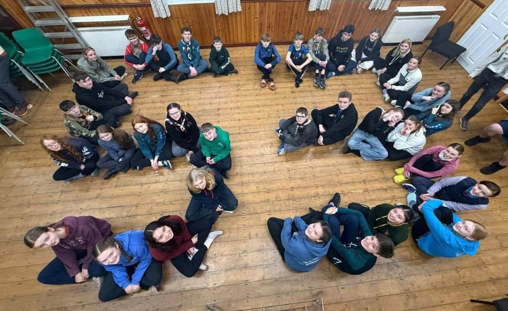 Photo showing the 36 young people who are members of the unit. The photo has been taking looking down on the group who are sat on a wooden floor and they form the shape of the number 53.