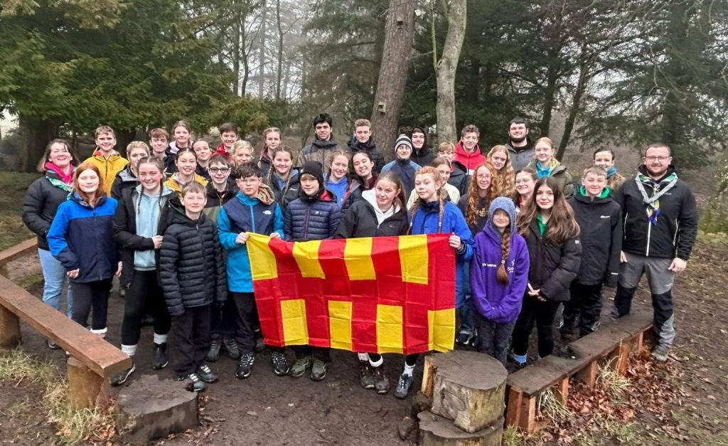 Group photo of the young people and leaders of Unit 53 standing outdoors, with trees in the background. Members of the group are holding a Northumberland flag.